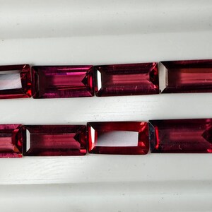 May include: Eight rectangular, faceted gemstones in a deep red colour are arranged in two rows. The gems have a polished appearance, reflecting light. The stones are set against a white background, highlighting their colour and shape. These gems are suitable for jewellery making.