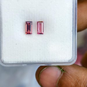 May include: Two rectangular, pink gemstones are displayed in a clear plastic case. The gems have a rectangular cut and a vibrant pink hue. The case is held by a person's hand, showcasing the gems' color and shape.