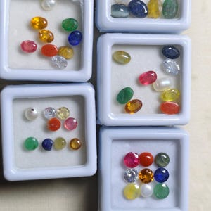 Puede incluir: Una colección de piedras preciosas de varios colores, incluyendo rojo, amarillo, verde, azul y transparente, exhibidas en pequeños contenedores cuadrados blancos. Las piedras preciosas son ovaladas, redondas y facetadas.