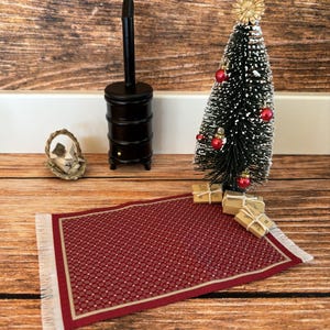 Könnte beinhalten: Miniatur-Weihnachtsszene mit einem kleinen schwarzen Weihnachtsbaum, geschmückt mit roten Kugeln und einem goldenen Stern. Ein roter Teppich mit weißem Fransen, ein schwarzer Ofen und verpackte Geschenke sind auf einer Holzoberfläche angeordnet.
