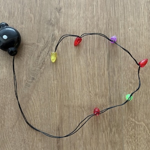 Könnte beinhalten: Eine Lichterkette mit bunten Weihnachtslichtern und einem schwarzen Batteriepack. Die Lichter sind rot, gelb, grün und lila. Die Lichter befinden sich an einem schwarzen Draht. Der Batteriepack ist schwarz und rund.