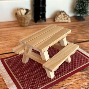 Könnte beinhalten: Ein Miniatur-Picknicktisch aus Holz mit angebrachten Bänken. Der Tisch ist hellbraun und steht auf einem rot gemusterten Teppich mit weißem Fransen. Die Szene umfasst einen kleinen schwarzen Ofen, einen Korb und einen Miniatur-Weihnachtsbaum.