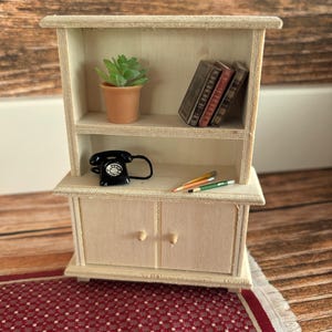 Könnte beinhalten: Eine weiße Holz-Hütte mit zwei Türen und zwei Regalen. Das obere Regal hat eine kleine Topfpflanze und einen Stapel Bücher. Das untere Regal hat ein schwarzes Drehtelefon und drei Bleistifte. Die Hütte steht auf einem roten und weißen gemusterten Teppich.