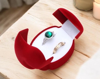 May include: A red velvet ring box with two rings inside. One ring has a green gemstone and the other is a plain band.