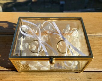 May include: A rectangular glass ring box with a gold-colored metal frame. Inside, two gold rings are secured with white ribbons to a bed of white flowers. The box is closed with a small gold knob.