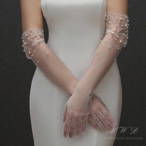 Könnte beinhalten: Transparente, elfenbeinfarbene Handschuhe mit verstreuten, runden Perlen. Die Handschuhe sind lang, reichen über die Ellbogen hinaus und bestehen aus einem zarten, durchsichtigen Stoff. Ein elegantes Design für formelle Anlässe.