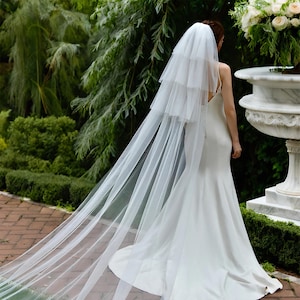 Puede incluir: Una novia con un vestido de novia blanco y velo está de pie al aire libre. El velo es largo y en capas, cayendo por la espalda del vestido. El vestido es de diseño sencillo y elegante, con un corpiño ajustado y una falda fluida. El fondo presenta vegetación y una fuente decorativa.