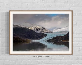 May include: A panoramic view of a glacier in a mountainous landscape. The glacier is a bright blue and white color, and the mountains are covered in snow and trees. The water in the foreground is calm and reflective.