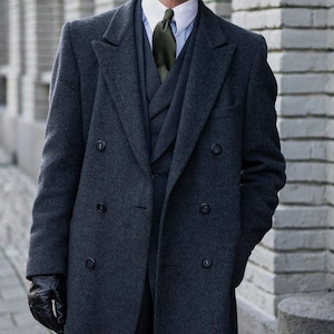 May include: A dark gray double-breasted overcoat with a black leather glove on the left hand. The person is wearing a dark suit with a white shirt and a dark green tie.