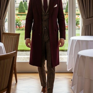 May include: A burgundy overcoat worn over a matching suit and tie. The suit includes a vest and plaid trousers. The outfit is completed with brown leather shoes. The setting appears to be a formal dining room with a view of a garden.