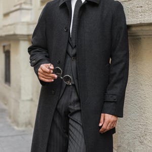 May include: A charcoal gray overcoat with a classic design. The coat features a notched collar, button closures, and a tailored fit. The person is wearing a white shirt, a patterned tie, and pinstripe pants. The person is holding a pair of sunglasses.