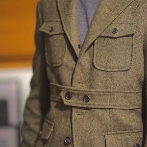 Może przedstawiać: Brązowa kurtka z tweedu w jodełkę z wieloma kieszeniami i paskiem w talii. Kurtka ma klasyczny krój z czarnymi guzikami i klapami z wycięciami. Modelka ma na sobie szary golf.