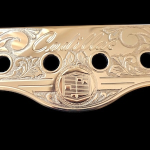 Puede incluir: Un emblema Cadillac de metal dorado con el nombre de la marca grabado en cursiva. El emblema presenta cuatro agujeros circulares y adornos florales ornamentados. El logotipo de Cadillac está centrado.