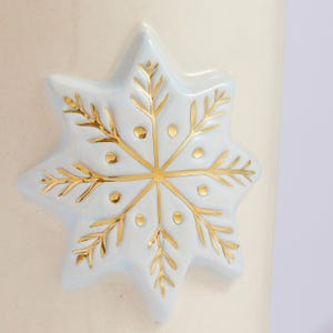 Puede incluir: Una taza de cer&aacute;mica blanca con un dise&ntilde;o de copo de nieve azul. El copo de nieve est&aacute; bordeado de oro y tiene peque&ntilde;os puntos dorados.