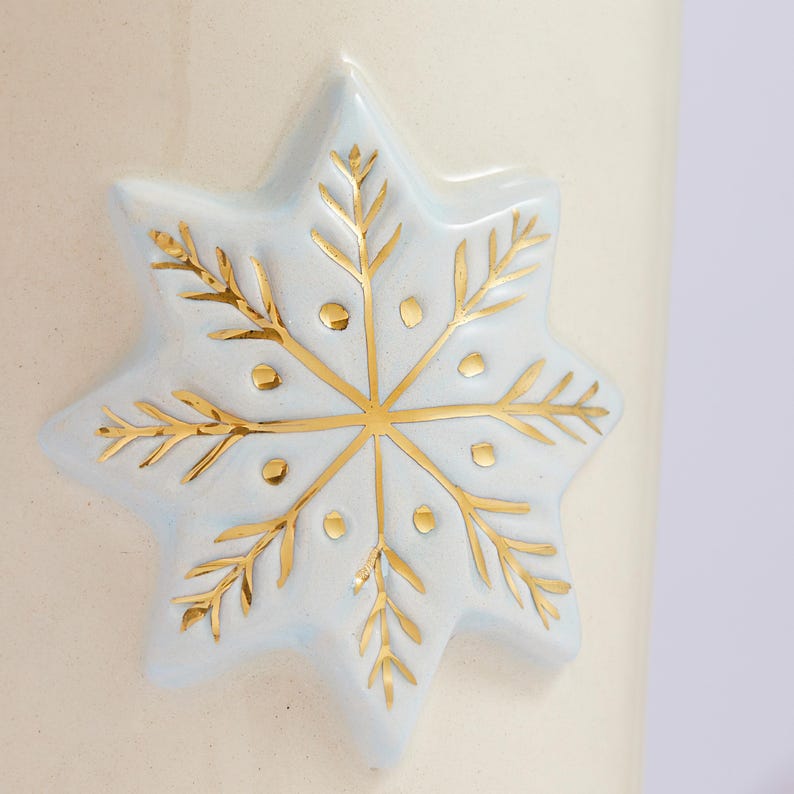 Puede incluir: Una taza de cer&aacute;mica blanca con un dise&ntilde;o de copo de nieve azul. El copo de nieve est&aacute; bordeado de oro y tiene peque&ntilde;os puntos dorados.