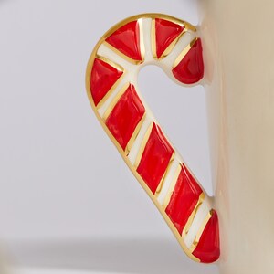 Puede incluir: Una taza de cer&aacute;mica blanca con un asa de bast&oacute;n de caramelo rojo y blanco. El asa est&aacute; decorada con detalles dorados.