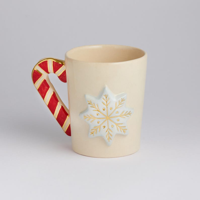 Puede incluir: Una taza de cer&aacute;mica blanca con un asa de bast&oacute;n de caramelo y un dise&ntilde;o de copo de nieve en relieve en oro y azul.