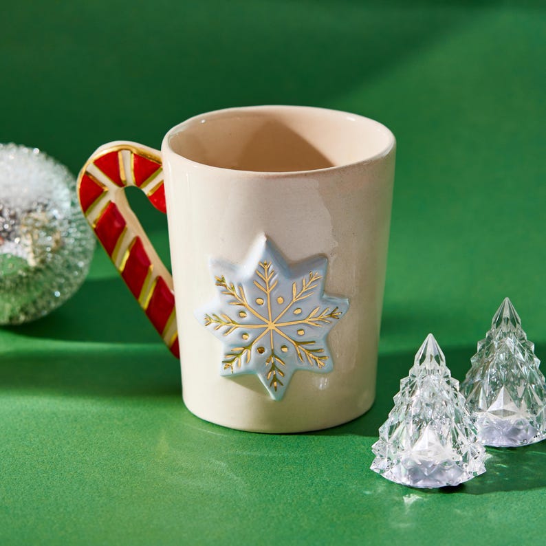 Puede incluir: Una taza de cer&aacute;mica blanca con un asa de bast&oacute;n de caramelo y un dise&ntilde;o de copo de nieve azul. La taza est&aacute; sobre una superficie verde con dos &aacute;rboles de Navidad de cristal transparente.