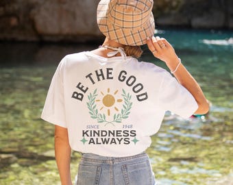 May include: White t-shirt with a graphic print of a sun with a laurel wreath and the text "Be the Good Since 1948 Kindness Always".