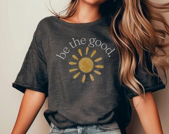 May include: A dark gray t-shirt with a yellow sun graphic and the text "be the good." printed in white.