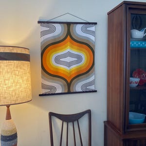 Puede incluir: Un tapiz de pared vintage con un diseño geométrico en tonos naranja, amarillo, marrón y blanco. El tapiz está colgado en una pared con un marco de madera.