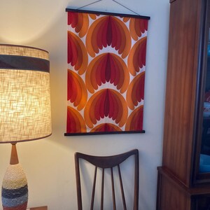 Op de afbeelding: Een vintage wandtapijt met een geometrisch patroon in tinten oranje, rood en wit. Het tapijt is ingelijst met een houten lat en hangt aan een witte muur.