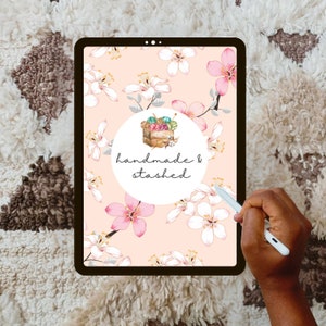 May include: A digital tablet displaying a pink and white floral pattern with a white circle in the center. The circle contains a drawing of a basket filled with yarn balls and knitting needles. The text "handmade & stashed" is written below the basket.