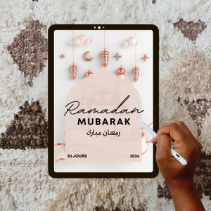 May include: A digital tablet displaying a graphic for Ramadan Mubarak. The graphic features a pink background with a crescent moon, stars, and lanterns. The text "Ramadan Mubarak" is written in black with the Arabic translation below. The text "30 jours" and "2024" are also included.