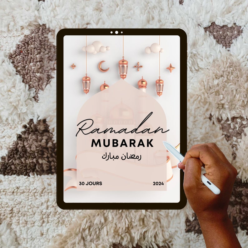 May include: A digital tablet displaying a graphic for Ramadan Mubarak. The graphic features a pink background with a crescent moon, stars, and lanterns. The text "Ramadan Mubarak" is written in black with the Arabic translation below. The text "30 jours" and "2024" are also included.