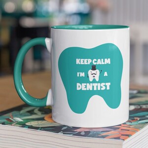 Könnte beinhalten: Eine weiße Keramiktasse mit einem türkisfarbenen Rand und Henkel. Die Tasse hat ein türkisfarbenes Zahnmotiv mit einem schwarzen Zylinder und einem Schnurrbart. Der Text "Keep Calm I'm A Dentist" ist auf den Zahn gedruckt.