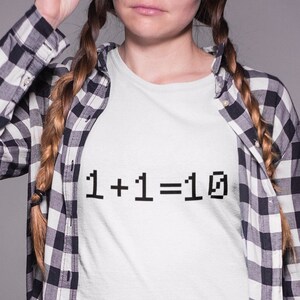 May include: A white t-shirt with black text that reads "1+1=10". The person wearing the shirt is also wearing a black and white checked shirt and brown trousers.