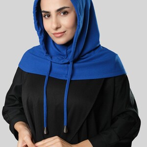May include: A black and blue hooded garment. The hood is a vibrant blue with drawstrings and silver-toned metal aglets. The garment has long sleeves and a black body. The model is wearing the garment.