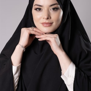 May include: A woman wearing a black hijab and a matching black abaya. The abaya is draped over a white collared shirt. She is wearing a silver bracelet and ring. The background is a solid gray.