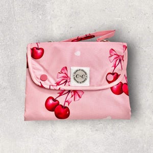 May include: A pink baby changing mat with a cherry print and a handle. The mat has a repeating pattern of red cherries and pink bows on a light pink background. The brand name "Cookie & Cherkie" is on a small label.