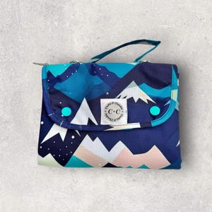 May include: A foldable changing mat featuring a mountain landscape pattern in blues, teals, and white. It has a handle and fastens with two turquoise buttons. A white label with the text "Cookie & Cheeks C & C" is centered on the front of the mat.