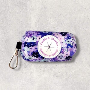 May include: Purple and white floral print dog poo bag dispenser with a silver clip and a round white label with the text "Cookie & Chawkie" in pink.