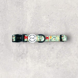 May include: A dog collar featuring a repeating pattern of different dog breeds on a pale green background. The collar has a black plastic buckle and a metal D-ring. A white circular tag with the text "Cookie & Chewbies" and "C&C" is attached.
