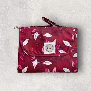 May include: A burgundy changing mat with a floral design of white and pink leaves. The mat has a small handle and a snap closure. A white label with the text "Cookie & Cheeks C+C" is centered on the front. The mat is folded.