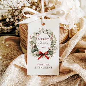 May include: A rectangular white gift tag featuring a watercolor wreath of green leaves, red berries, and cotton bolls. The text reads Merry Christmas 2026 and With Love, The Greens. A white ribbon is tied through the top hole of the paper tag.