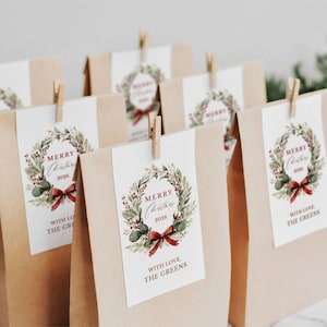 May include: A group of brown paper bags, each featuring a white rectangular tag attached with a wooden clip. The tags display a watercolor Christmas wreath illustration, the text Merry Christmas 2026, and the phrase With Love, The Greens.