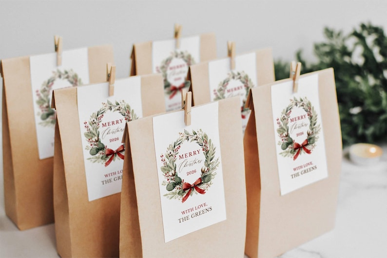 May include: A group of brown paper bags, each featuring a white rectangular tag attached with a wooden clip. The tags display a watercolor Christmas wreath illustration, the text Merry Christmas 2026, and the phrase With Love, The Greens.