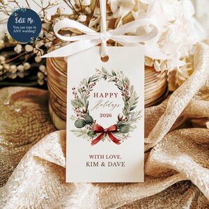 May include: A rectangular white gift tag featuring a watercolor wreath of green leaves, red berries, and cotton bolls. The text reads Happy Holidays 2026 and With Love, Kim & Dave. A white ribbon is tied through the top hole of the paper tag.