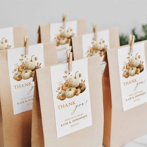 Puede incluir: Un grupo de bolsas de papel marrón, cada una con una etiqueta rectangular blanca sujeta con una pinza de madera. Las etiquetas muestran una ilustración en acuarela de calabazas y hojas de otoño con el texto "THANK you! WITH LOVE FROM KATIE & DOMINIQUE".
