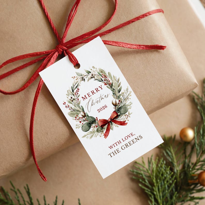 May include: A white rectangular gift tag featuring a watercolor wreath of green leaves, red berries, and cotton bolls. The text reads Merry Christmas 2026 and With Love, The Greens. A red ribbon ties the tag to a brown paper wrapped gift.