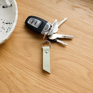 Op de afbeelding: Een lichtgroene leren sleutelhanger met de letters "LL" erop gegraveerd. De sleutelhanger is bevestigd aan een set sleutels, waaronder een autosleutel met het Audi-logo.
