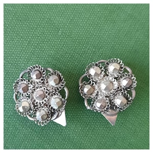 Op de afbeelding: Twee zilverkleurige metalen sluitingen met een bloemmotief. Elke sluiting heeft een cluster van facetgeslepen, zilverkleurige stenen omgeven door een decoratief, opengewerkt frame. De sluitingen hebben een driehoekige bevestiging aan de onderkant.