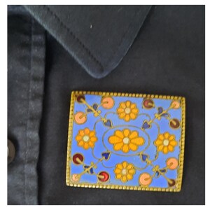 Op de afbeelding: Vierkante broche met een blauwe achtergrond en een gouden rand. De broche heeft een bloemenontwerp met oranje en gele bloemen, roze cirkels en blauwe hartvormige accenten. De broche is geplaatst op een zwarte stof.