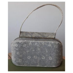 Op de afbeelding: Een vintage handtas met een textuur, grijs en wit patroon. De rechthoekige tas heeft een gebogen handvat en een metalen sluiting. Het ontwerp van de tas heeft een herhalend patroon van kleine, lichtgekleurde vormen.