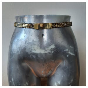 Op de afbeelding: Een goud-zwarte kettingriem met een rechthoekige gouden gesp met een zonnesterontwerp. De riem wordt getoond op een zilveren mannequin torso tegen een witte achtergrond. Het is een modeaccessoire.