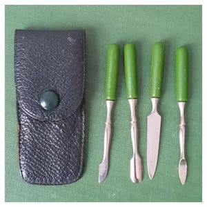 Könnte beinhalten: Ein Vintage-Maniküre-Set mit einem schwarzen Lederetui und vier Werkzeugen mit grünen Griffen. Die Werkzeuge umfassen einen Nagelhautschieber, einen Löffel, eine Nagelfeile und ein gebogenes Werkzeug. Das Set wird auf einer grünen Oberfläche präsentiert.